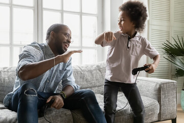 Wall Mural - Happy african American dad and little son play video games enjoy family weekend at home together, overjoyed biracial father and small ethnic boy child engaged in funny leisure entertainment activity