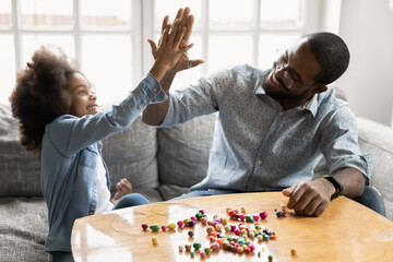 Overjoyed young biracial father and little daughter give high five celebrate achievement make bracelet together, happy african American dad and small girl child have fun do creative activity together