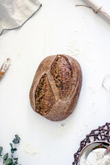 A loaf of country sourdough bread on a light table next to flour, napkins, sprigs of plants, , top view, vertical background, copy space, flay lay, top-down food