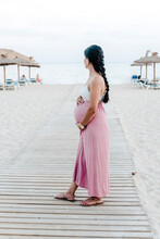 Young Pregnant Latin Woman On An Almost Empty Beach On A Wood Walkway, Looking At The Horizon With A Long Braid. Feeling Of Peace And Tranquility On A Lovely Summer Day.