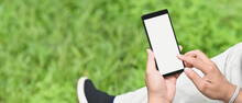 A Man Is Using A White Blank Screen Smartphone While Sitting Over The Grass Field As A Background.