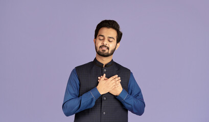 Portrait of Indian guy with closed eyes holding his hands near heart on lilac background