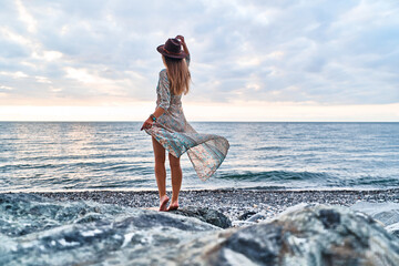 Boho chic woman in long fluttering dress and felt hat standing back on stone by the sea