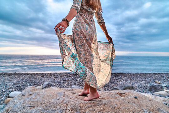 Boho woman with long waving dress standing on a stone by the seashore at sunset