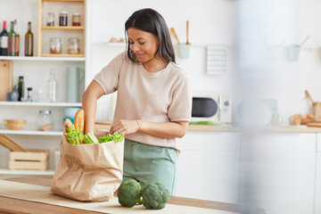 Waist up portrait of elegant mixed-race woman unpacking bags with food while standing in white kitchen interior and smiling, food delivery service, copy space