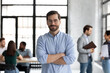 © fizkes - Head shot portrait smiling confident businessman wearing glasses standing in modern office room with arms crossed, diverse colleagues on background, executive boss startup founder looking at camera