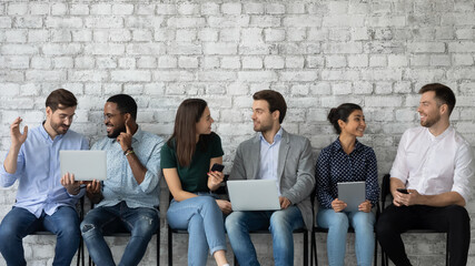 Friendly diverse candidates talking, chatting and using gadgets while waiting for job interview, young business people applicants sitting in row in queue, recruitment and employment concept