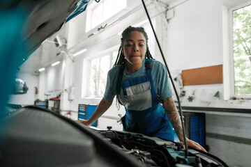 Wall Mural - True care. Young african american woman, professional female mechanic looking, examining under hood of car at auto repair shop