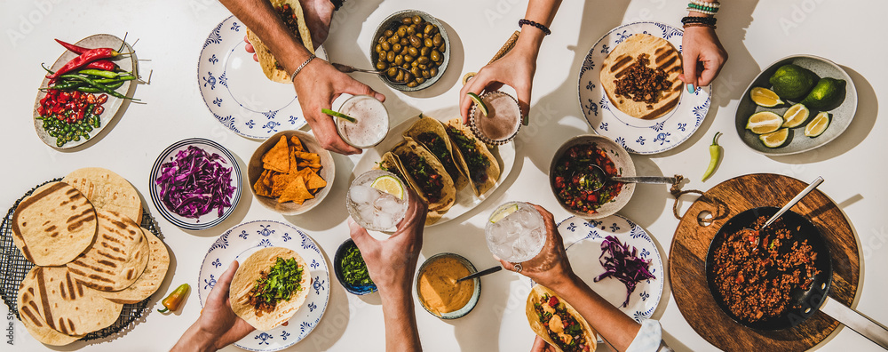 Friends having Mexican Taco dinner. Flat-lay of beef tacos, tomato salsa, tortillas, beer, snacks and peoples hands clinking glasses over white table, top view. Mexican cuisine, comfort food concept - obrazy, fototapety, plakaty 
