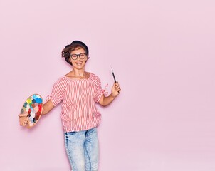 Young beautiful artist woman wearing glasses and beret smiling happy. Jumping with smile on face painting using paintbrush and palette over isolated pink background