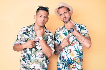 Young gay couple of two men wearing summer hat and hawaiian shirt in hurry pointing to watch time, impatience, looking at the camera with relaxed expression