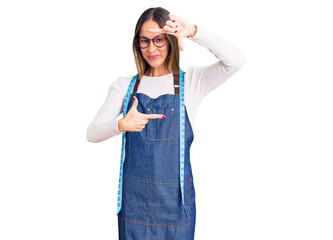 Beautiful brunette young woman dressmaker designer wearing atelier apron smiling making frame with hands and fingers with happy face. creativity and photography concept.