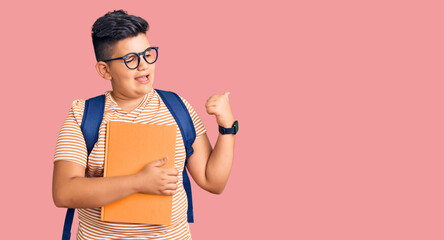 Little boy kid wearing student backpack holding books pointing thumb up to the side smiling happy with open mouth