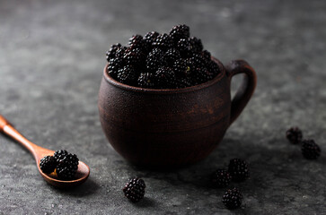 Blackberries in an earthenware mug are piled with a hill. Next to it is a wooden spoon in which the berries are scattered around on the table