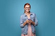 © Strelciuc - Red haired woman with eyeglasses smiling at camera while chatting on mobile posing on a blue studio wall