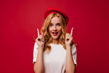 Ecstatic White Girl In Beret Posing With Amazement. Elegant Caucasian Female Model In T-shirt Standing On Red Background.