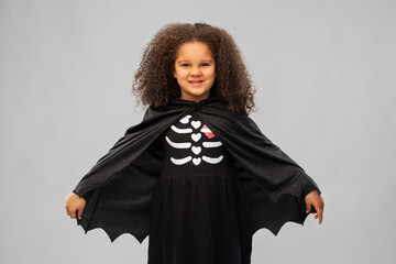 halloween, holiday and childhood concept - smiling african american girl in black dracula costume with bat's cape over grey background