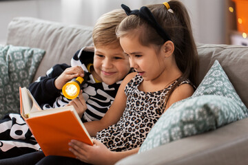 Poster - halloween, holiday and childhood concept - little kids in party costumes with flashlight reading book at home