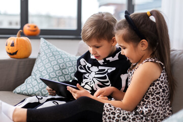 Wall Mural - halloween, holiday and childhood concept - smiling little boy and girl in party costumes with tablet pc computer at home