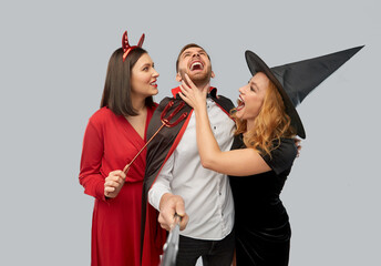 Poster - friendship, holiday and people concept - group of happy smiling friends in halloween costumes of witch, devil and vampire having fun taking picture by selfie stick over grey background