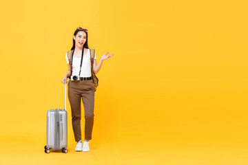 Wall Mural - Full length portrait of smiling cute young Asian woman tourist with trolley bag opening hand to copy space aside isolated on yellow studio background