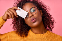 Portrait Of A Dark Skinned Curly Woman In Glasses And A Yellow Sweater With A Mockup Credit Card In Her Hands In A Photo Studio On A Pink Background. The Concept Of Easy Banking, Advertising.