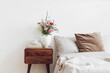 Cup of coffee and books on retro wooden bedside table. Rustic white ceramic vase with bouquet of pink cocmos and zinnia flowers. Beige linen and velvet pillows in bed. Scandinavian interior, bedroom.