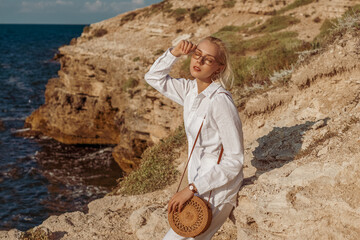 Outdoor summer fashion portrait of elegant woman wearing white linen suit, sunglasses, wrist watch, with round wicker bag, posing on rocks near sea. Copy, empty space for text