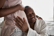© Roman - happy young african man is listening to the belly of his pregnant wife, couple in domestic wear at home