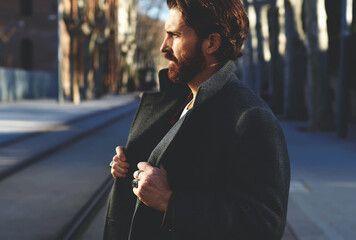 Portrait of fashionable well dressed man with beard posing outdoors looking away, confident and focused mature man in coat standing outside at sunny evening, elegant fashion model