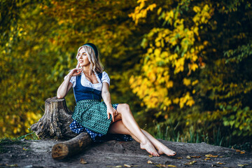 Wall Mural - Barefoot happy pretty blond girl in dirndl, traditional beer festival dress, sitting outdoors with blured colorful trees behind