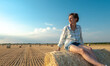 Stylish girl relaxing on hay bale in summer field in sunset. Young woman resting on hay on sunset, atmospheric tranquil moment. Countryside background