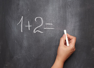 math example written in white chalk on a black chalk board and a woman's hand with white chalk