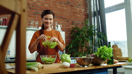Wall Mural - Woman preparing salad in kitchen. Smiling girl tearing lettuce on salad bowl.