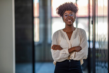 Wall Mural - Pretty young dreamy African-American office worker standing with arms crossed and looking at camera. Young business woman in modern office. Young African ethnicity businesswoman