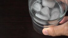 Place A Glass Of Ice Water On The Table. Man Putting On The Black Table A Glass With Ice Cubes And Fresh Water In Slow Motion. Top View.