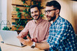 © BullRun - Portrait of cheerful handsome man looking at camera while his friend explaining information and idea  while cooperating, skilled male programmers working together on project using laptop computer
