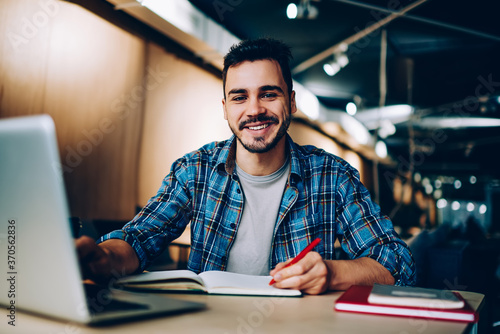 Cuadros en Lienzo Portrait of cheerful male student enjoying learning in coworking office using laptop computer for research,happy freelancer looking at camera during making project for remote job making notes