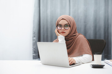 Wall Mural - Young muslim business woman wearing brown hijab working with laptop computer in her house. Look at camera.