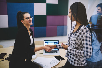 Young cheerful female colleagues shaking hands celebrating successful project and victory in competition, smiling business partners satisfied with agreement of collaboration planning work together