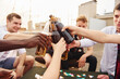 © standret - Doing cheers by bottles with beer. Group of young people in casual clothes have a party at rooftop together at daytime