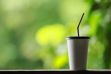 Wall Mural - Closeup of takeaway paper cup of iced coffee with plastic cover and straw on wooden table with green nature background and copy space.