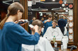 © Svitlana - Young professional barber girl working with hair clipper, serving young guy sitting in barber shop chair in front of the mirror. Selective focus