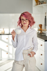 Smiling young stylish fashion hipster teen girl pink hair wears white shirt glasses holding cup drinking tea looking at camera standing in modern sunny home office kitchen interior. Vertical portrait.