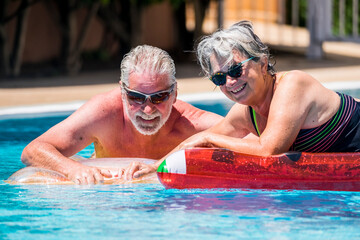 Wall Mural - Happy retired senior caucasian couple enjoy together the summer with an active lifestyle at the pool - people with age have fun with coloured inflatable lilos together