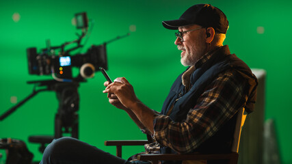 Wall Mural - Prominent Successful Director Sitting in a Chair on a Break Using Smartphone. On the Studio Film Set with High-End Equipment Professional Crew Shooting High Budget Movie.