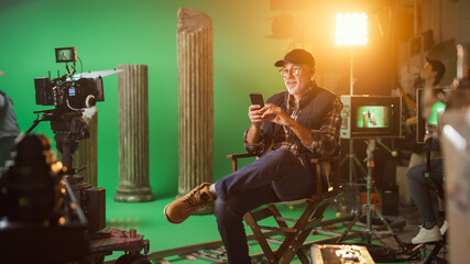 Wall Mural - Prominent Successful Director Sitting in a Chair on a Break Using Smartphone. On the Studio Film Set with High-End Equipment Professional Crew Shooting High Budget Movie.