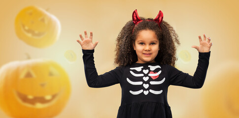 Wall Mural - halloween, holiday and childhood concept - smiling african american girl in black costume dress and red devil's horns over jack-o-lanterns on orange background