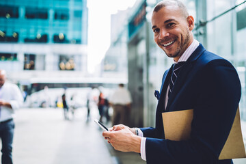 Portrait of cheerful trader satisfied with receiving money transaction getting notification on mobile standing near copy space area for advertising, successful businessman in formal wear sending sms.