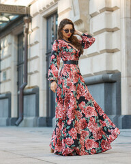 Young elegant lady in red and black floral design dress. Full length woman portrait. Beautiful girl standing and posing at city street. Fashionable female model with wavy hair